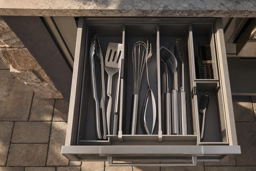 Outdoor kitchen cabinet drawer with adjustable dividers holding utensils and a meat thermometer in separate compartments